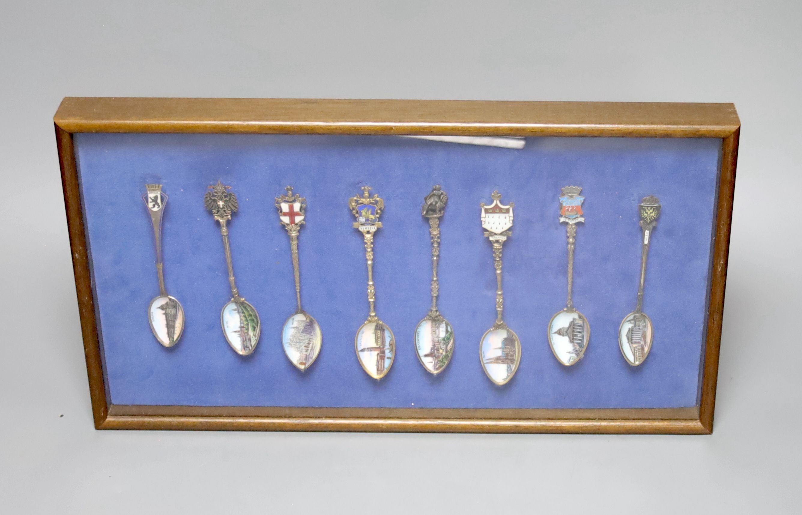 A collection of sixty four 800 standard silver (many gilt) and enamel souvenir spoons, early 20th century, in eight display cases, Including views of London, Germany, Italy, Austria, Spain etc.